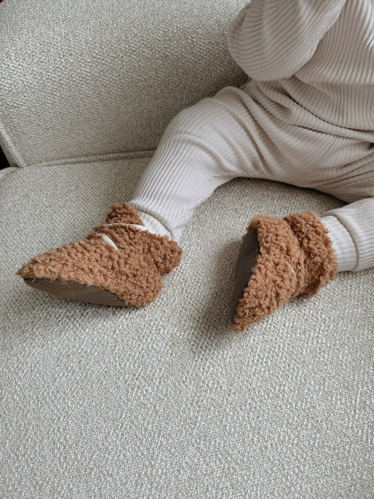 Close-up of brown teddy booties worn by a baby seated on a gray sofa.