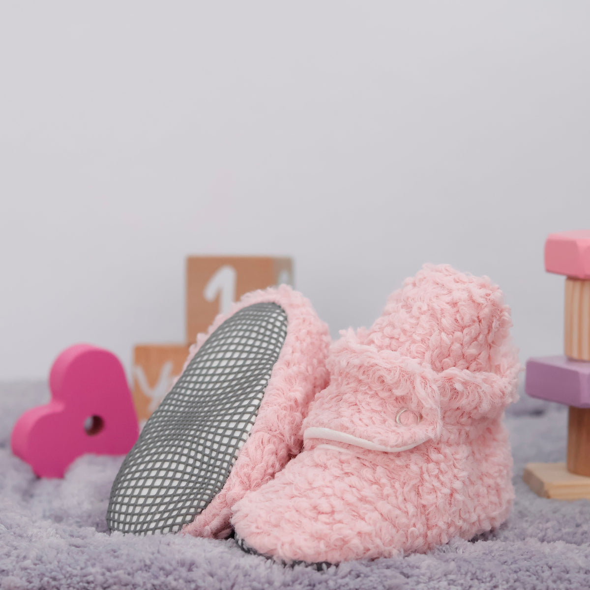 Pink teddy-texture baby booties with snap straps and gray grip soles arranged on a surface with one sole facing the camera.