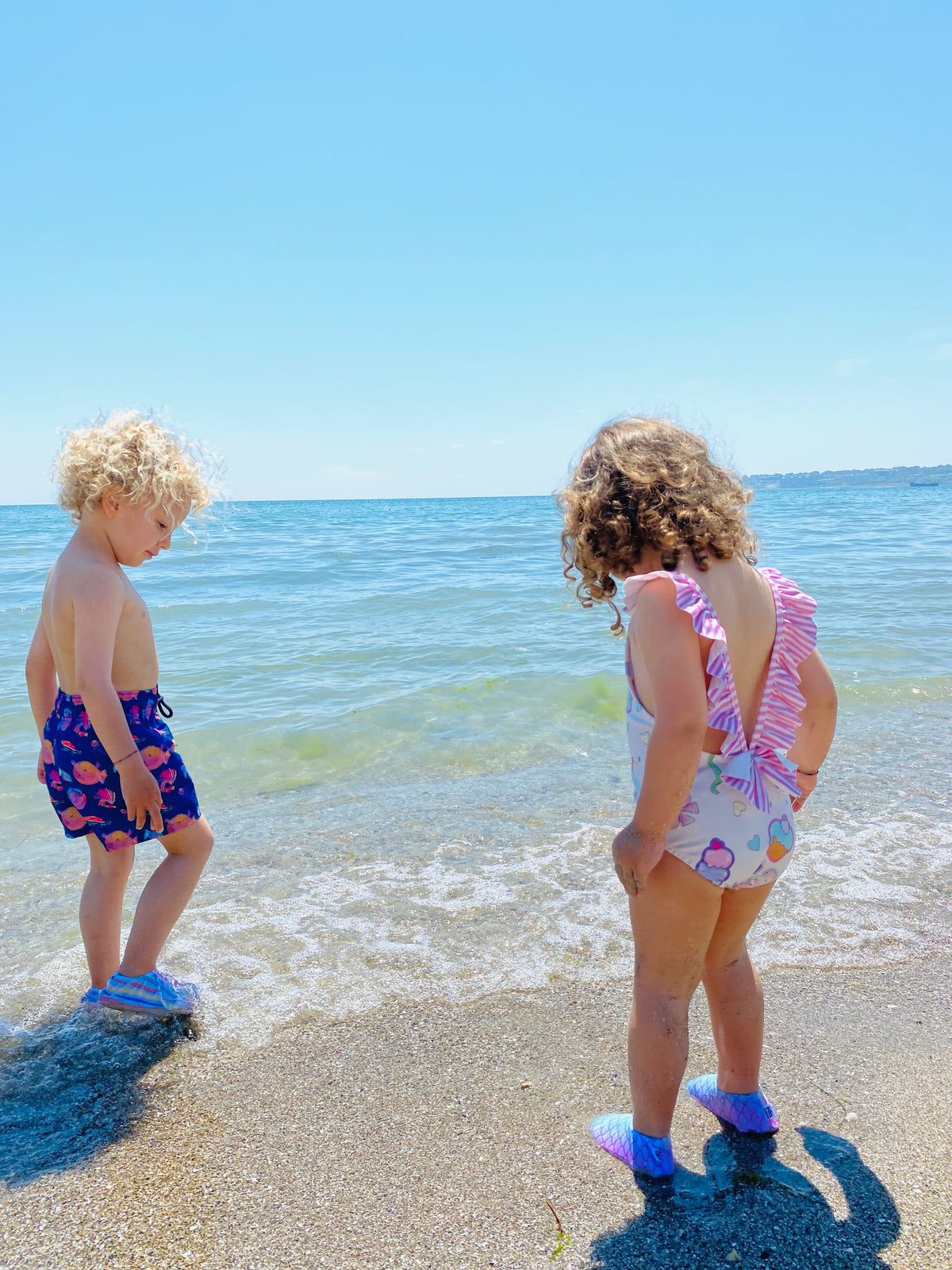 Two children standing at the shoreline wearing water shoes.