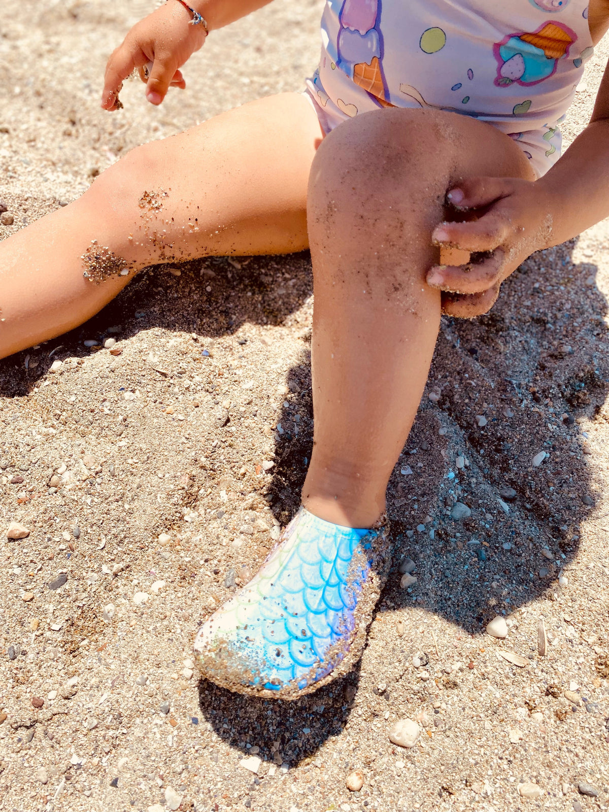 Rainbow scale-pattern water shoes worn by a child seated on a beach.