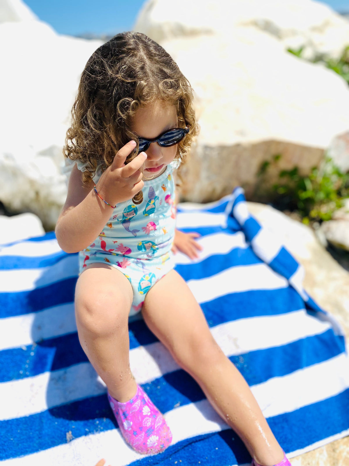 Child seated on a striped beach towel wearing pink shell-print water shoes.