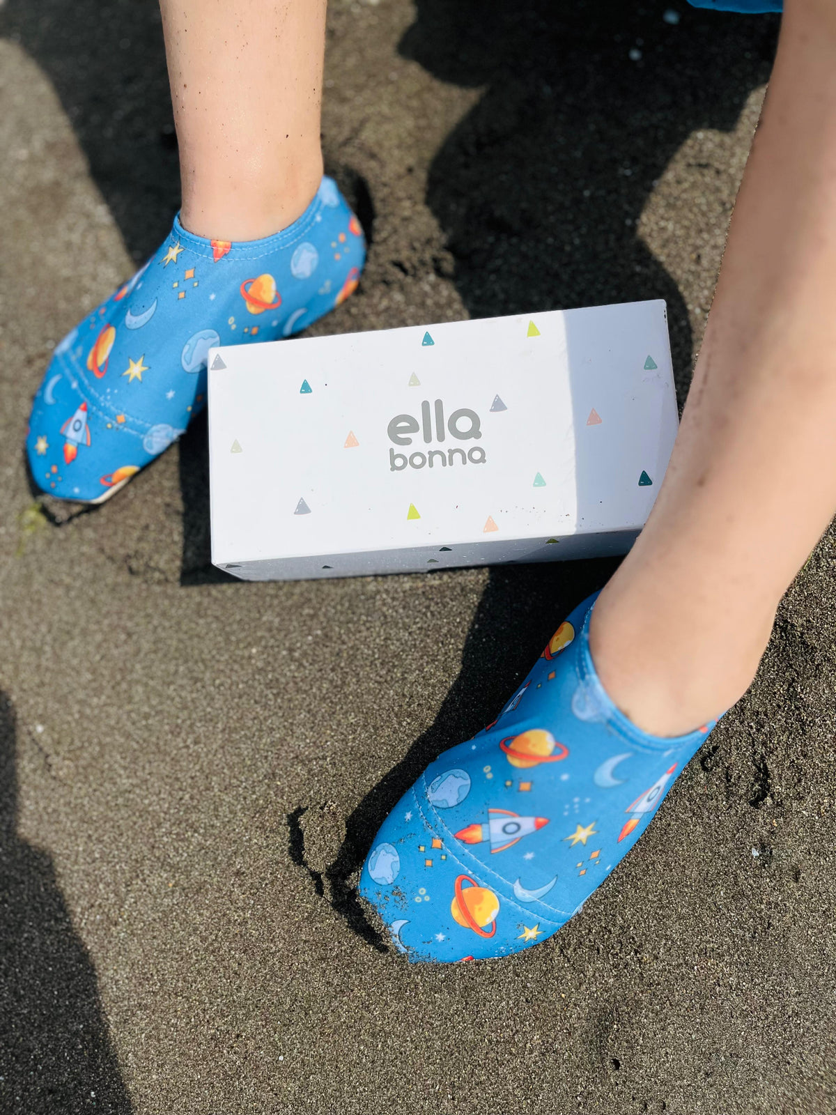 Close-up of space-print water shoes on wet sand beside an Ella Bonna card.