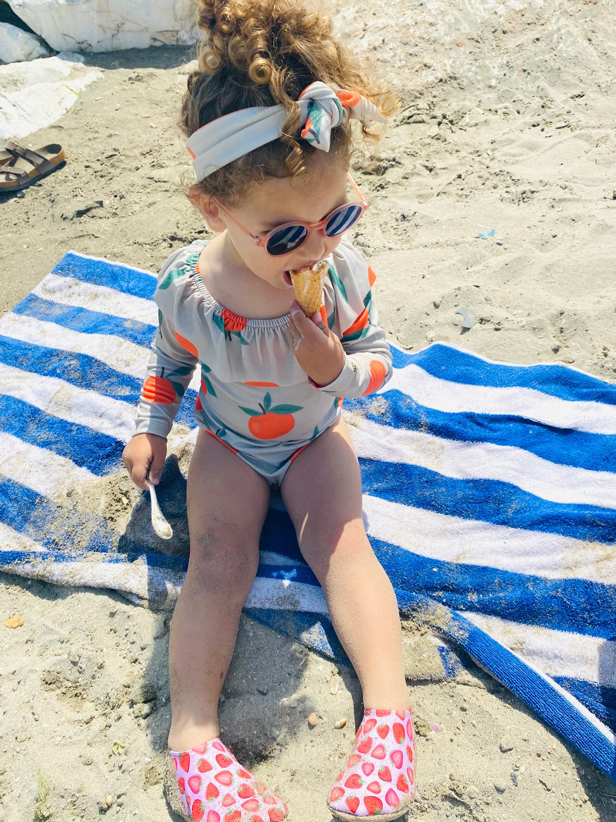 Child seated on a striped towel wearing strawberry-print water shoes.