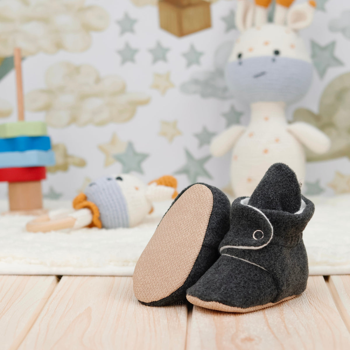 Charcoal fleece-texture baby booties with snap straps and tan soles arranged on a surface with one sole facing the camera.