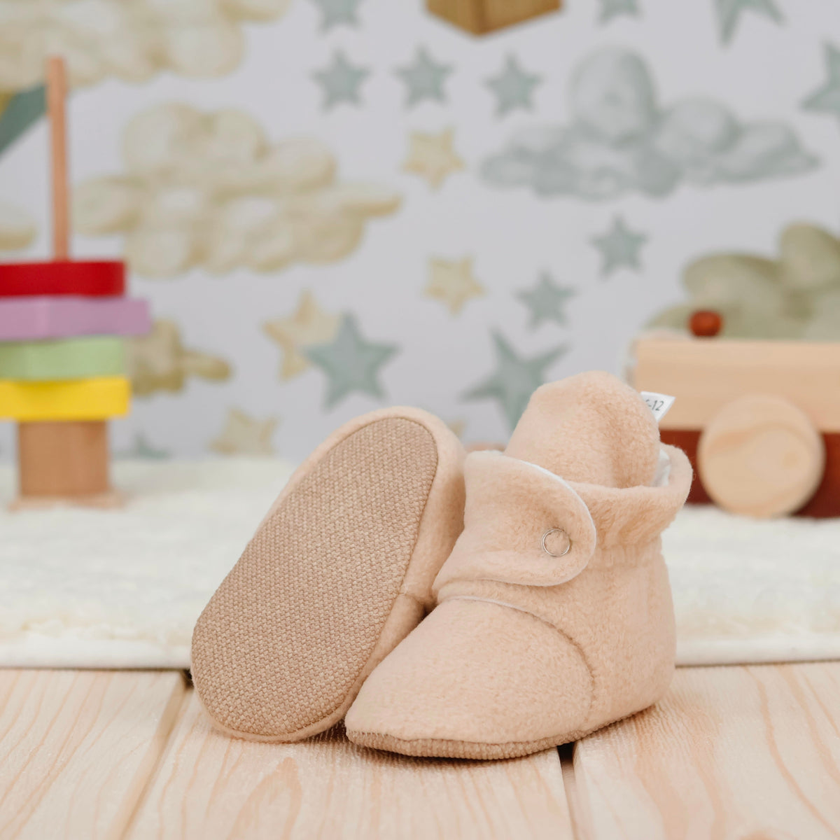Beige fleece-texture baby booties with snap straps and tan soles arranged on a surface with one sole facing the camera.