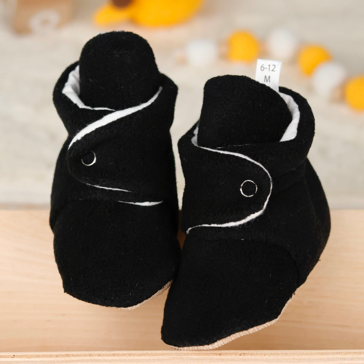 Front view of a pair of black fleece-texture baby booties with snap straps and tan soles.