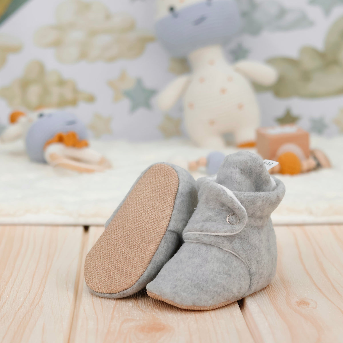 Light gray fleece-texture baby booties with snap straps and tan soles arranged on a surface with one sole facing the camera.