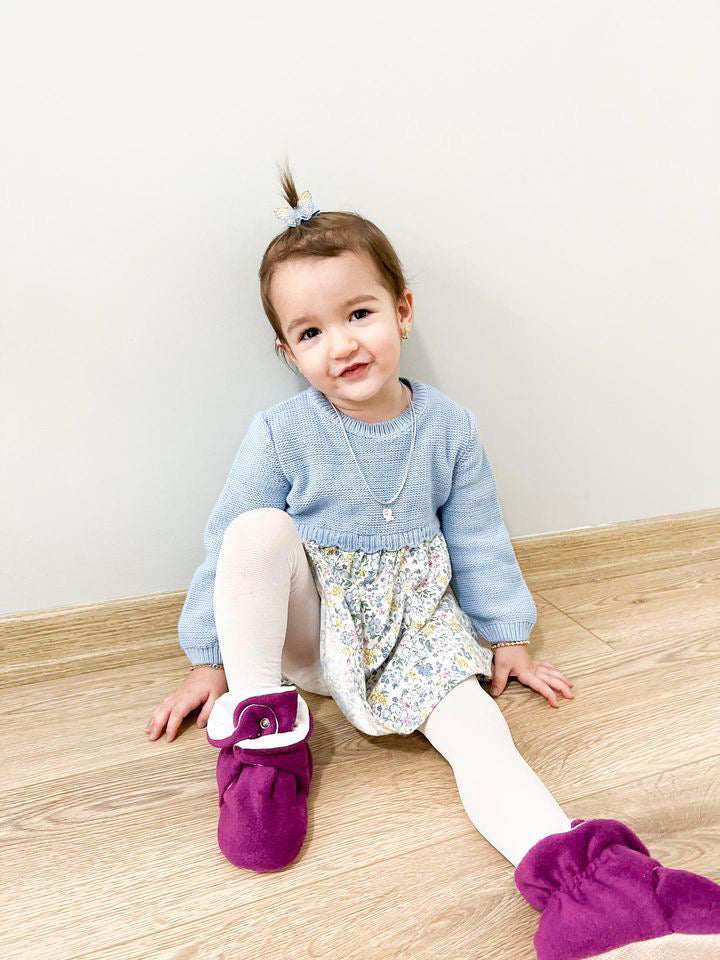 Child seated on a wooden floor wearing purple booties.