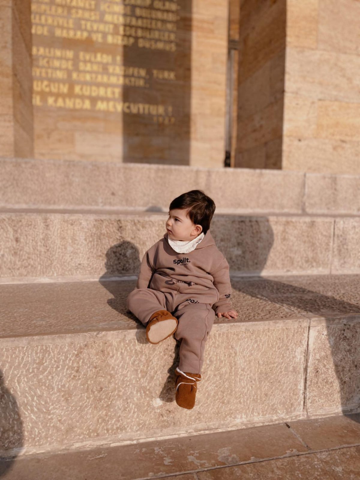 Child seated on a stone ledge wearing brown booties.