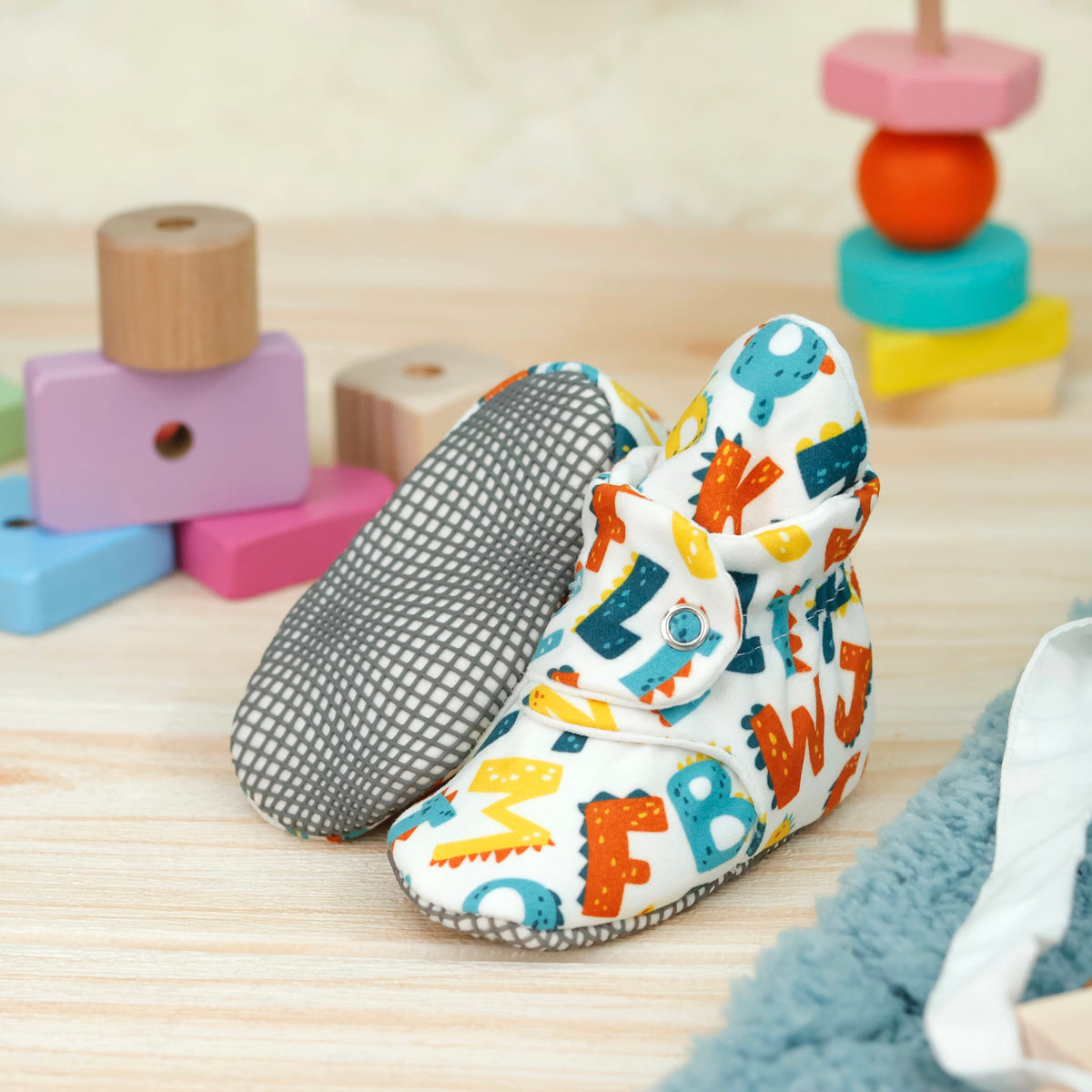 White fabric baby booties with colorful alphabet print and snap straps arranged on a surface with one sole facing the camera.