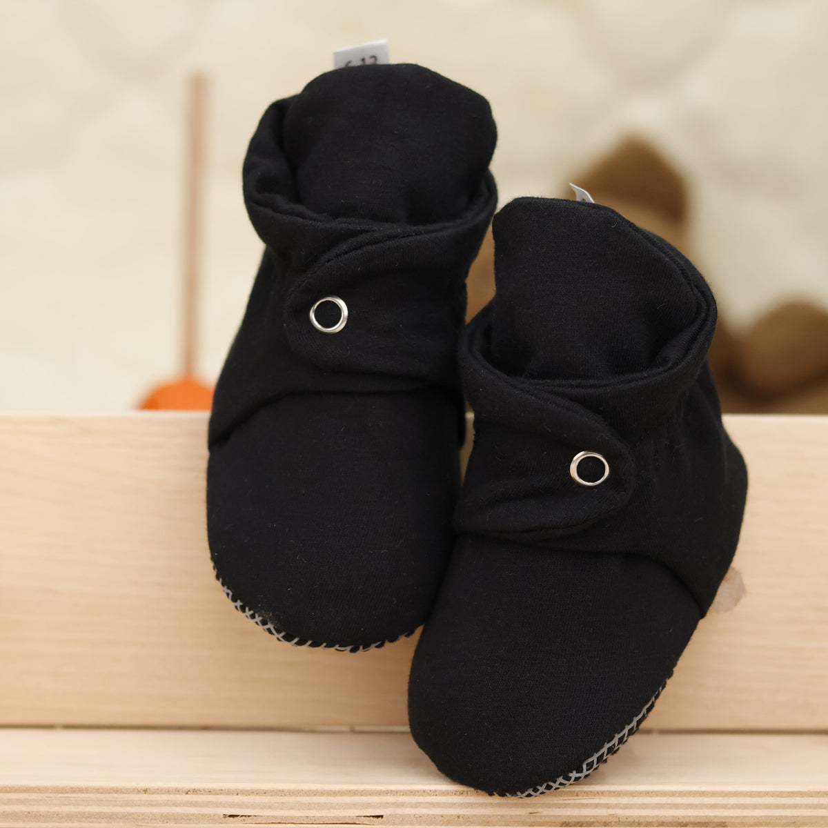 Front view of a pair of black fabric baby booties with snap straps and gray grip soles.