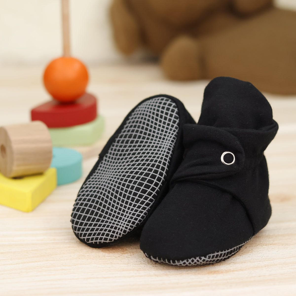 Black fabric baby booties with snap straps and gray grip soles arranged on a surface with one sole facing the camera.
