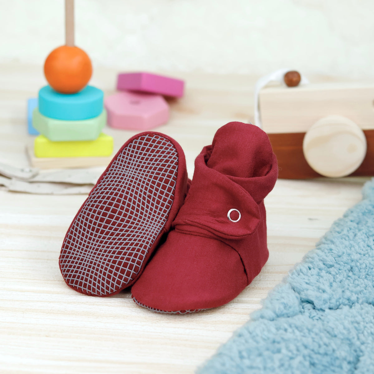 Burgundy fabric baby booties with snap straps and gray grip soles arranged on a surface with one sole facing the camera.