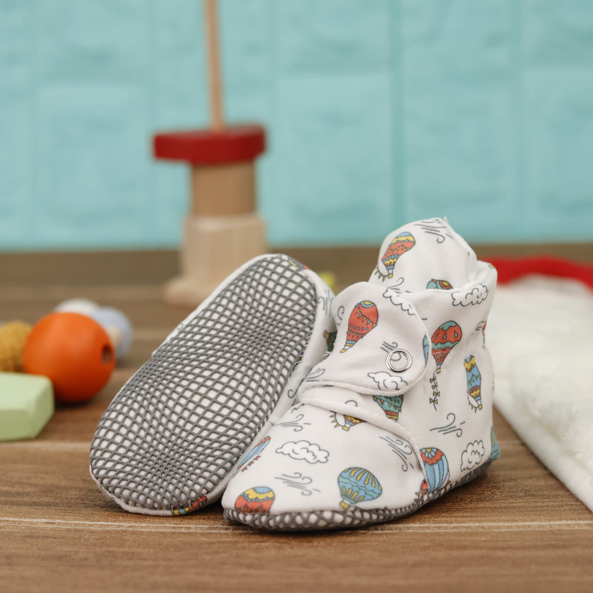 Cream baby booties with colorful hot-air balloons, clouds and snap straps arranged on a surface with one sole facing the camera.