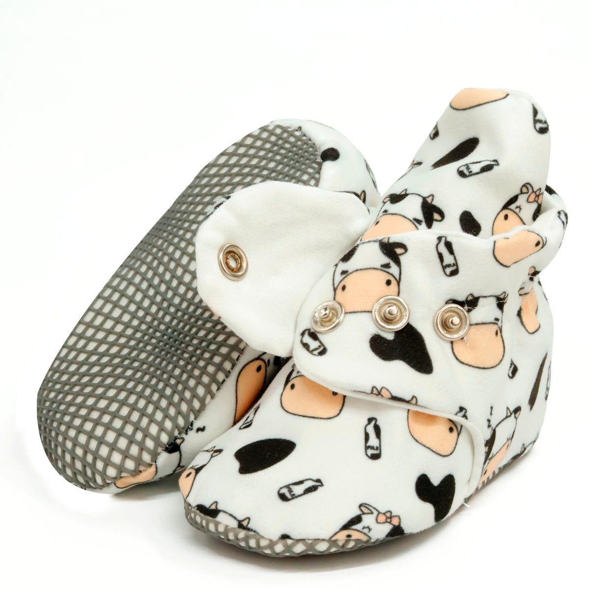 Cream baby booties with cartoon cows, black patches and snap straps, showing one sole and an open snap strap on white.