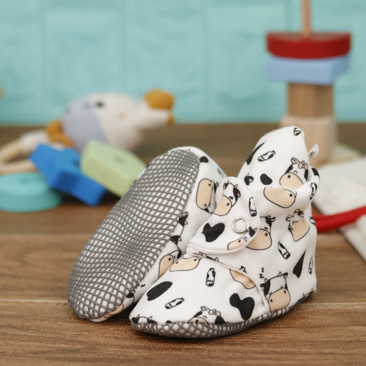 Cream baby booties with cartoon cows, black patches and snap straps arranged on a surface with one sole facing the camera.