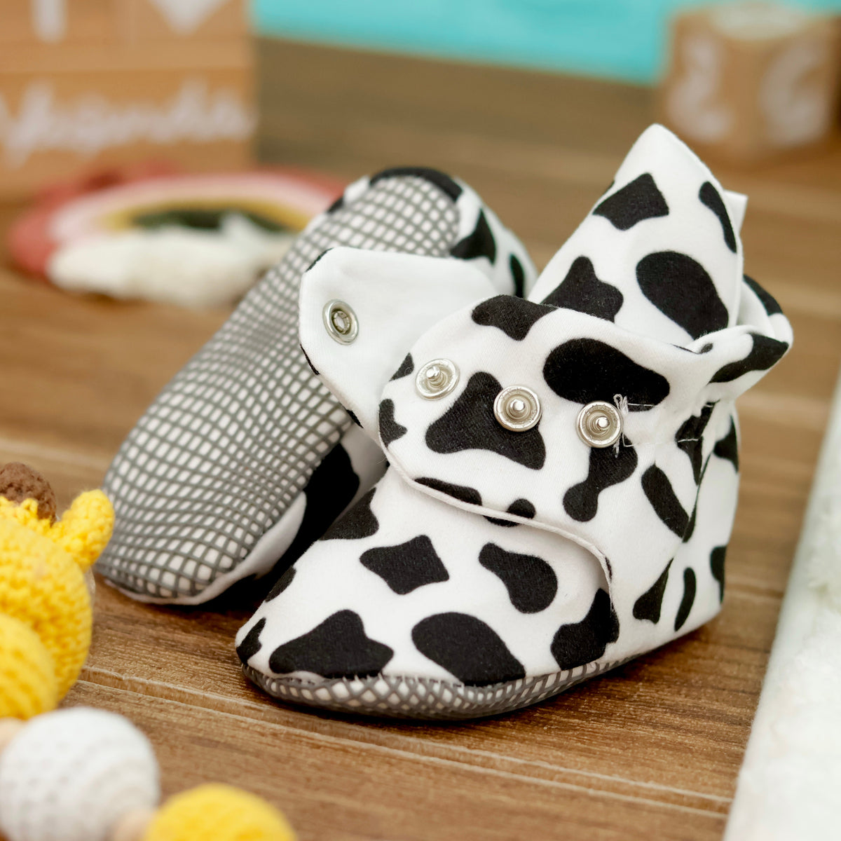 Close-up of white baby booties with black cow spots and snap straps, showing one sole and an open snap strap.