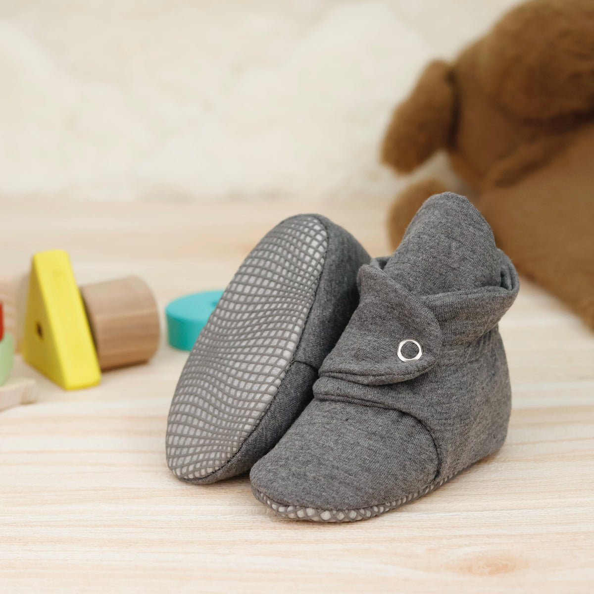 Dark gray fabric baby booties with snap straps and gray grip soles arranged on a surface with one sole facing the camera.