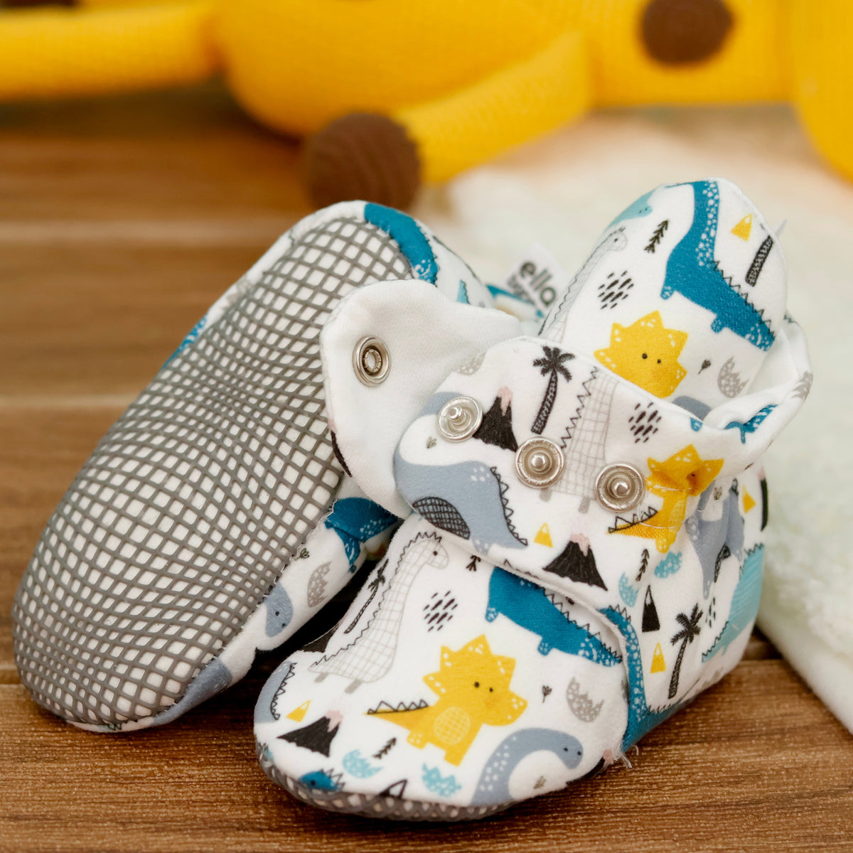 Close-up of cream baby booties with blue, gray and yellow dinosaurs and snap straps, showing one sole and an open snap strap.
