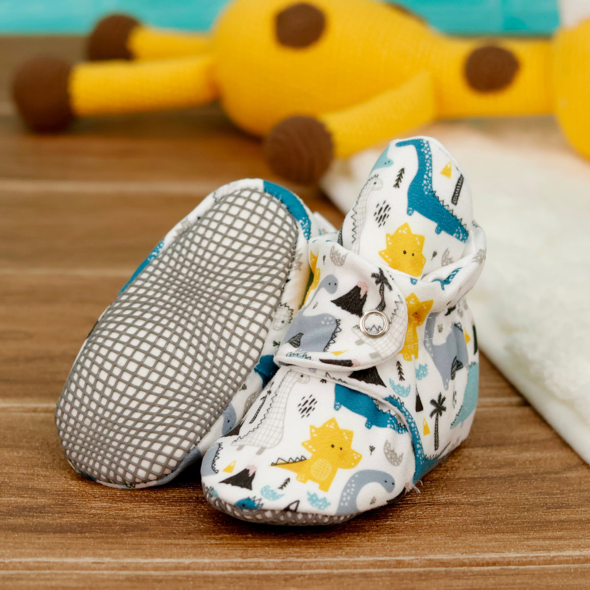 Cream baby booties with blue, gray and yellow dinosaurs and snap straps arranged on a surface with one sole facing the camera.