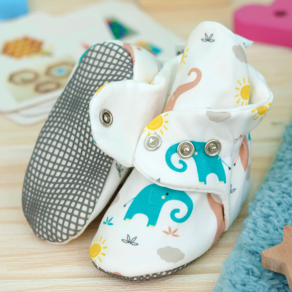 Close-up of cream baby booties with pastel elephant print and snap straps, showing one sole and an open snap strap.