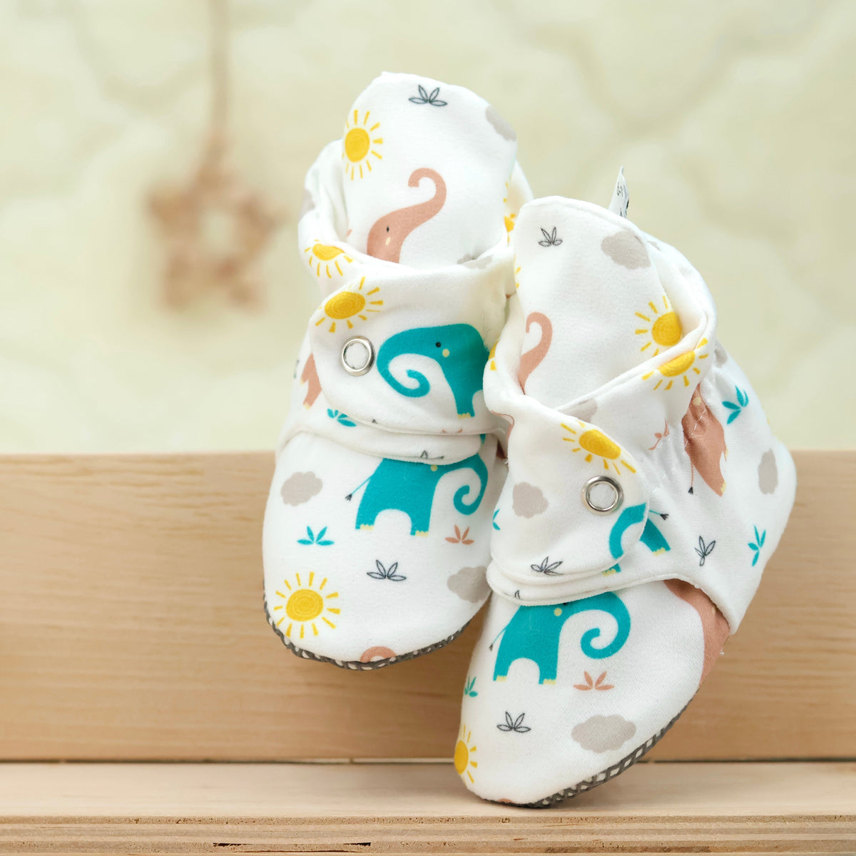 Front view of a pair of cream baby booties with pastel elephant print and snap straps.