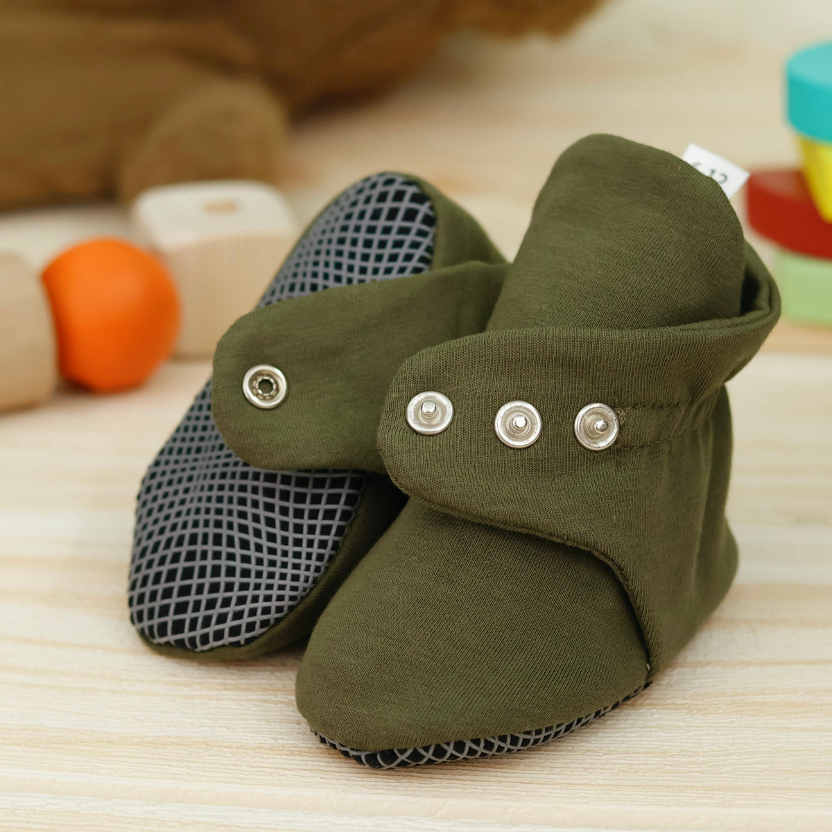 Close-up of olive green fabric baby booties with snap straps and gray grip soles, showing one sole and an open snap strap.