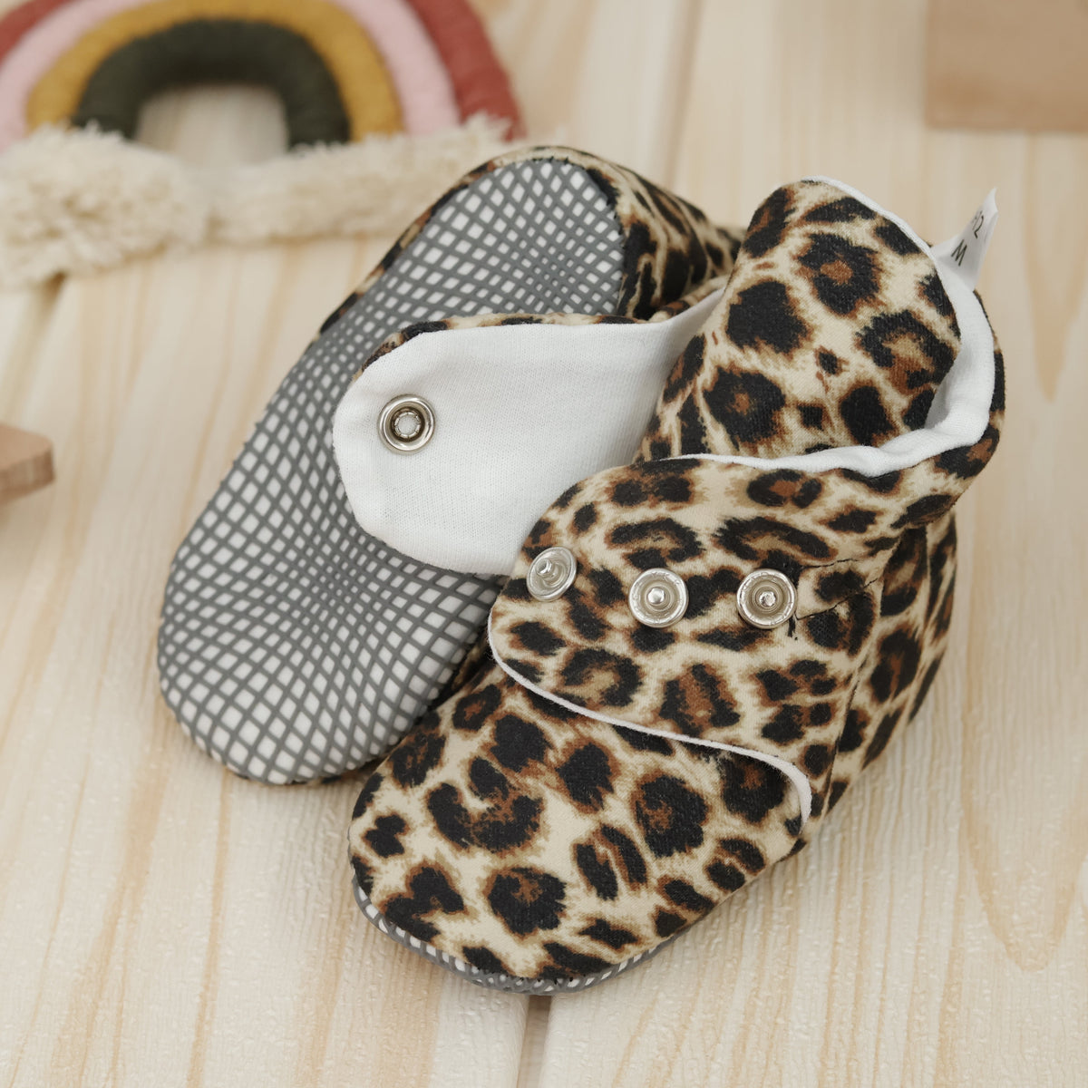 Close-up of tan baby booties with brown-and-black leopard spots and snap straps, showing one sole and an open snap strap.