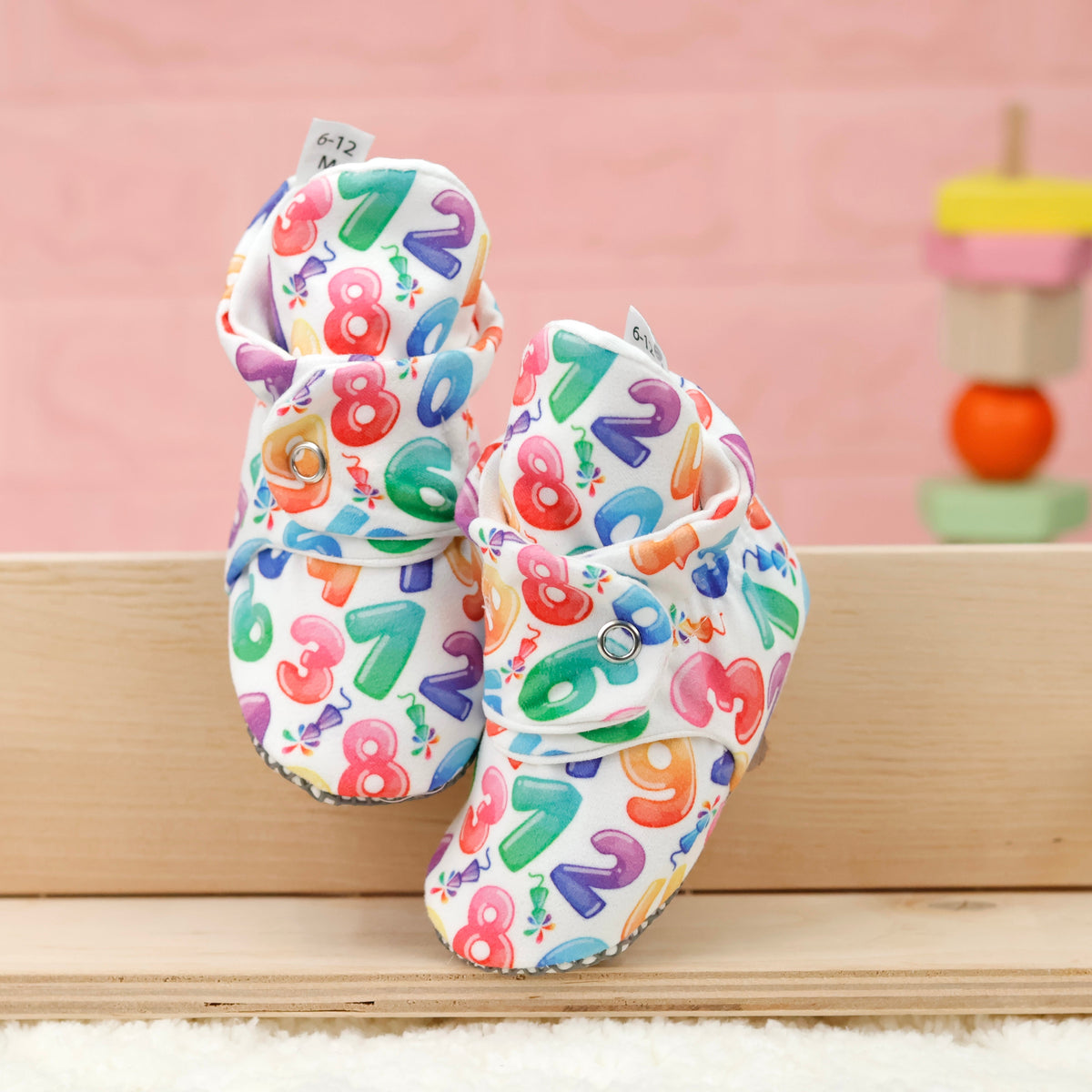 Front view of a pair of white baby booties with colorful numbers and snap straps.