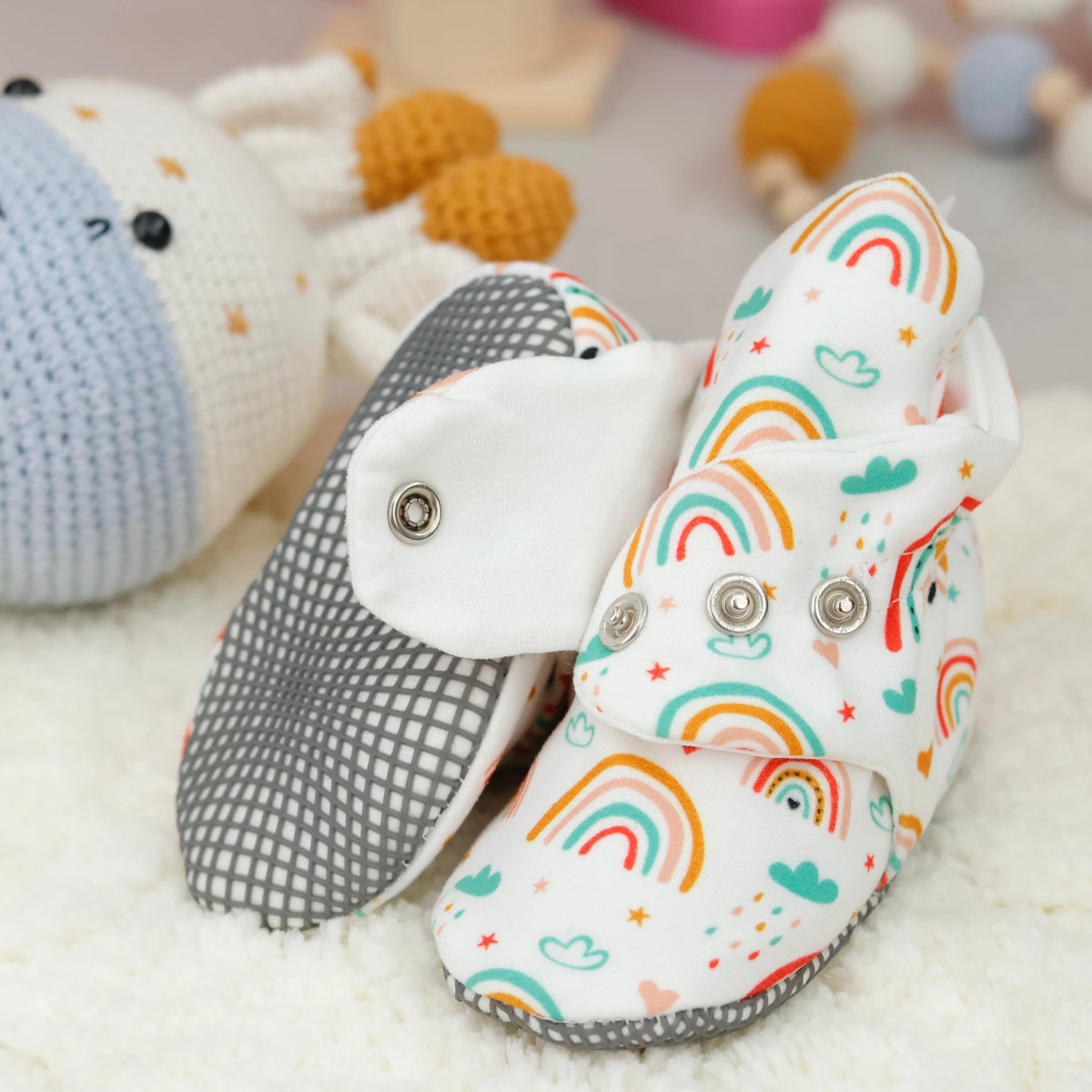 Close-up of cream baby booties with colorful rainbows and snap straps, showing one sole and an open snap strap.