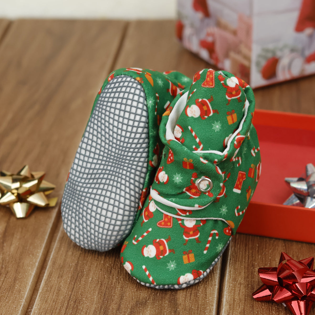 Green baby booties with Santa Claus Christmas print and snap straps arranged on a surface with one sole facing the camera.