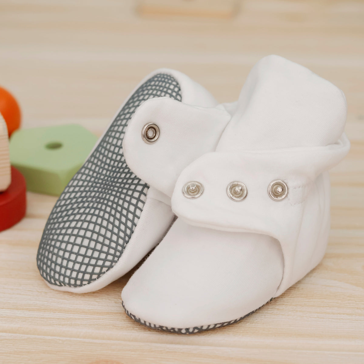 Close-up of white fabric baby booties with snap straps and gray grip soles, showing one sole and an open snap strap.