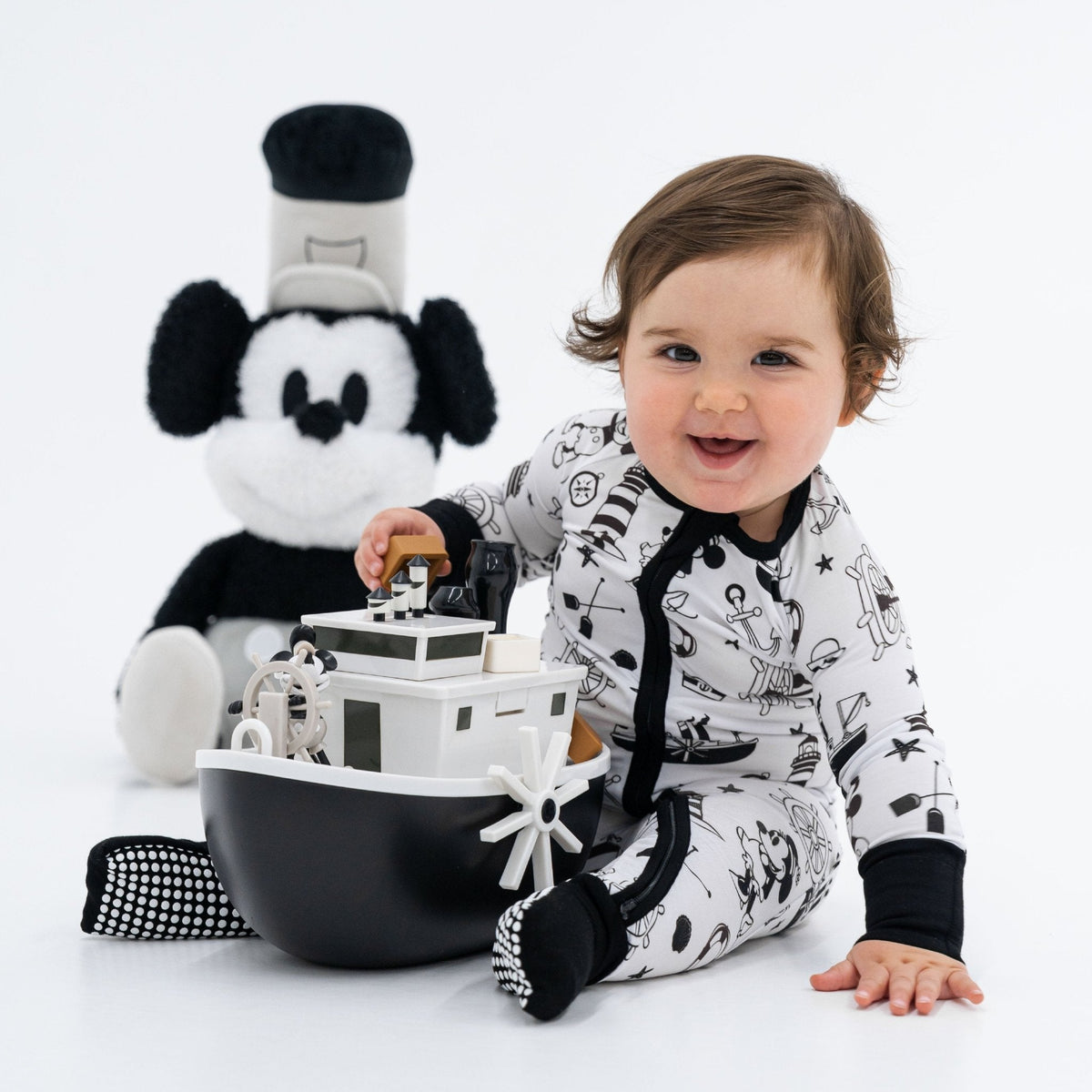 Baby sitting beside a toy boat while wearing Bam & Stitch Steamboat Willie Convertible Romper.