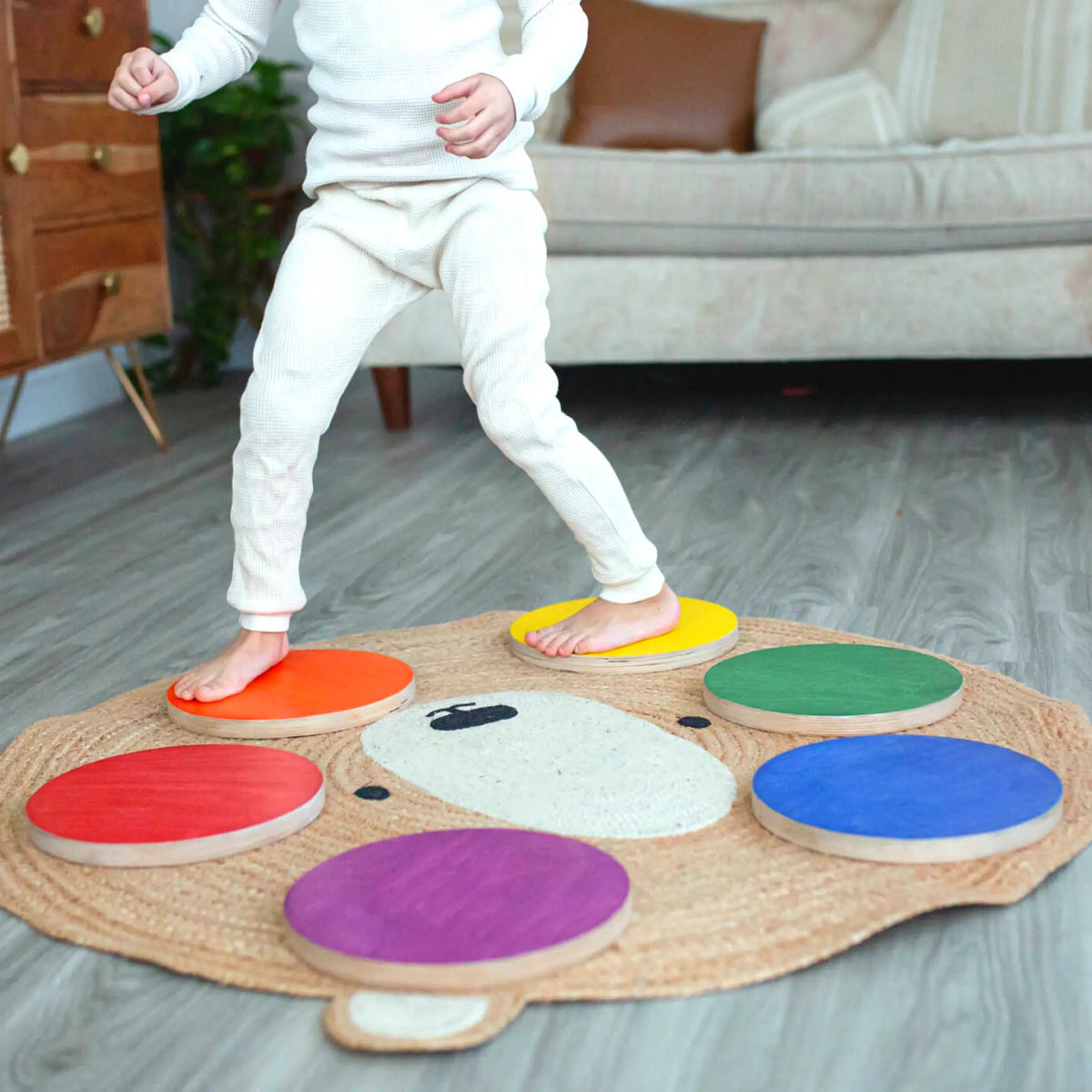 Child balancing between orange and yellow stepping stones with other rainbow stones on a bear-shaped rug.