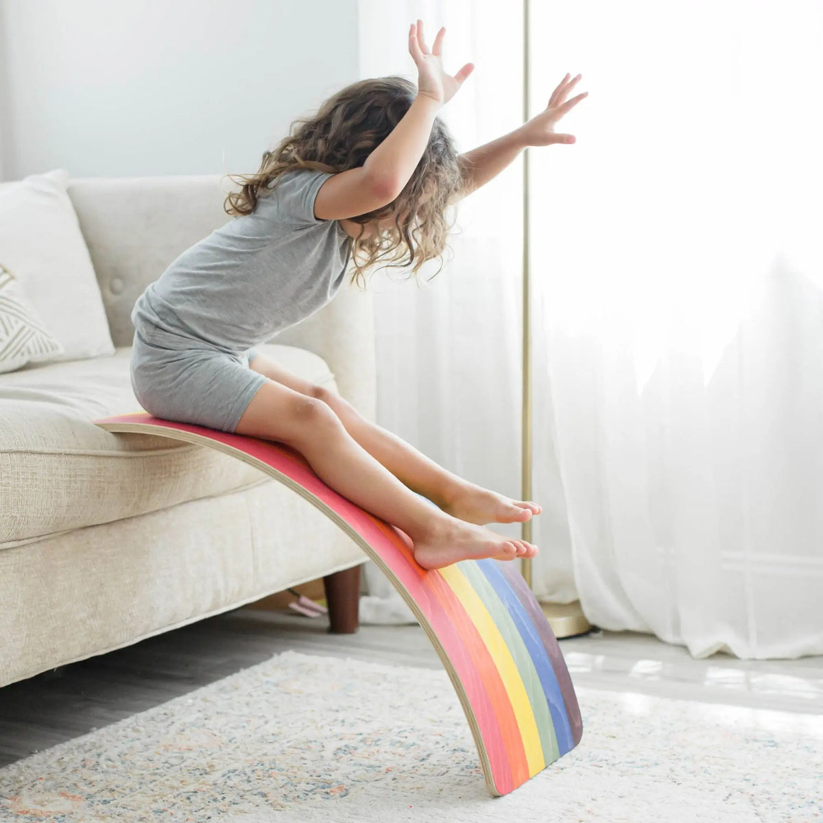 Child sitting on a rainbow-striped wobble board propped against a sofa as a ramp.