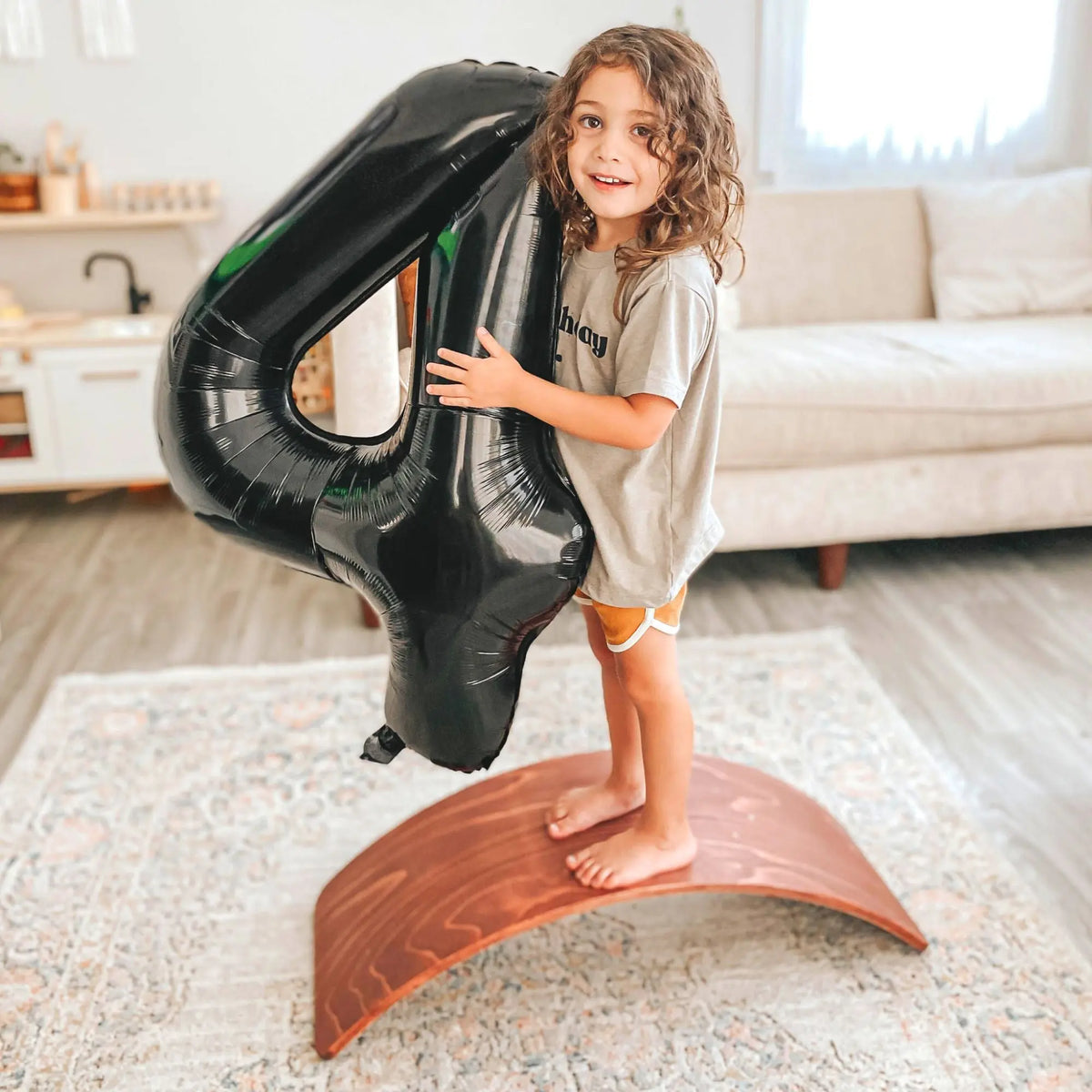 Child standing on the arch of a reddish-brown wobble board while holding a number-four balloon.
