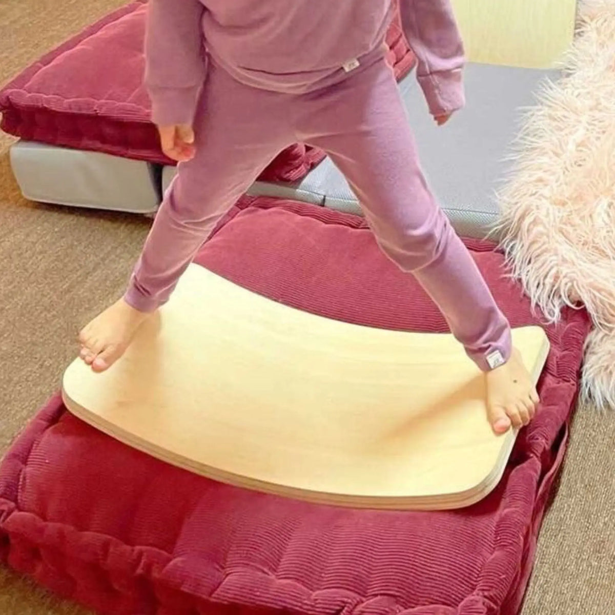 Child standing with feet apart on a pale wobble board placed on floor cushions.