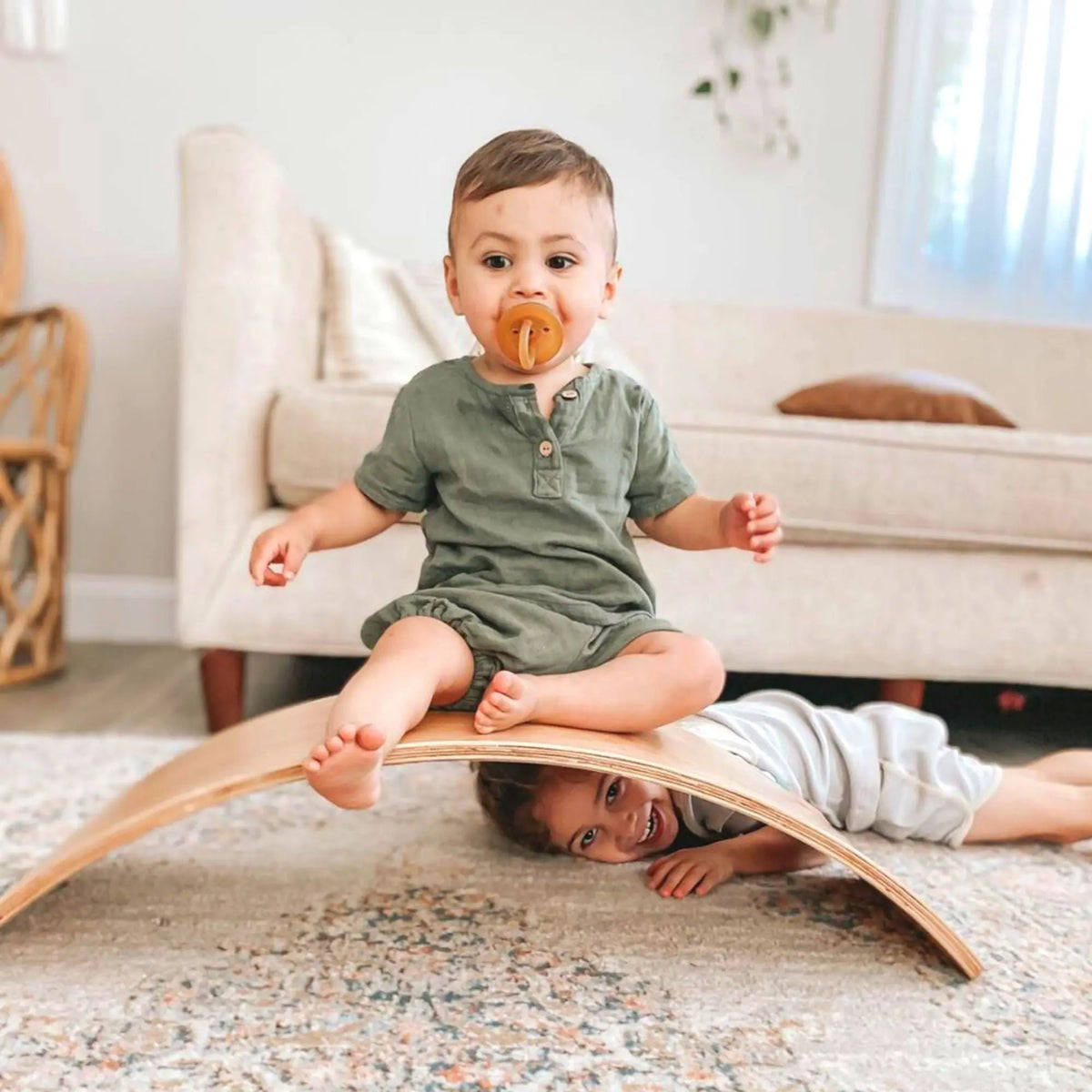 One child seated on the arch of a natural-tone wobble board while another peeks out from underneath.