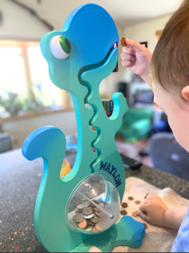 Child placing a coin in an aqua dinosaur bank showing personalized name lettering