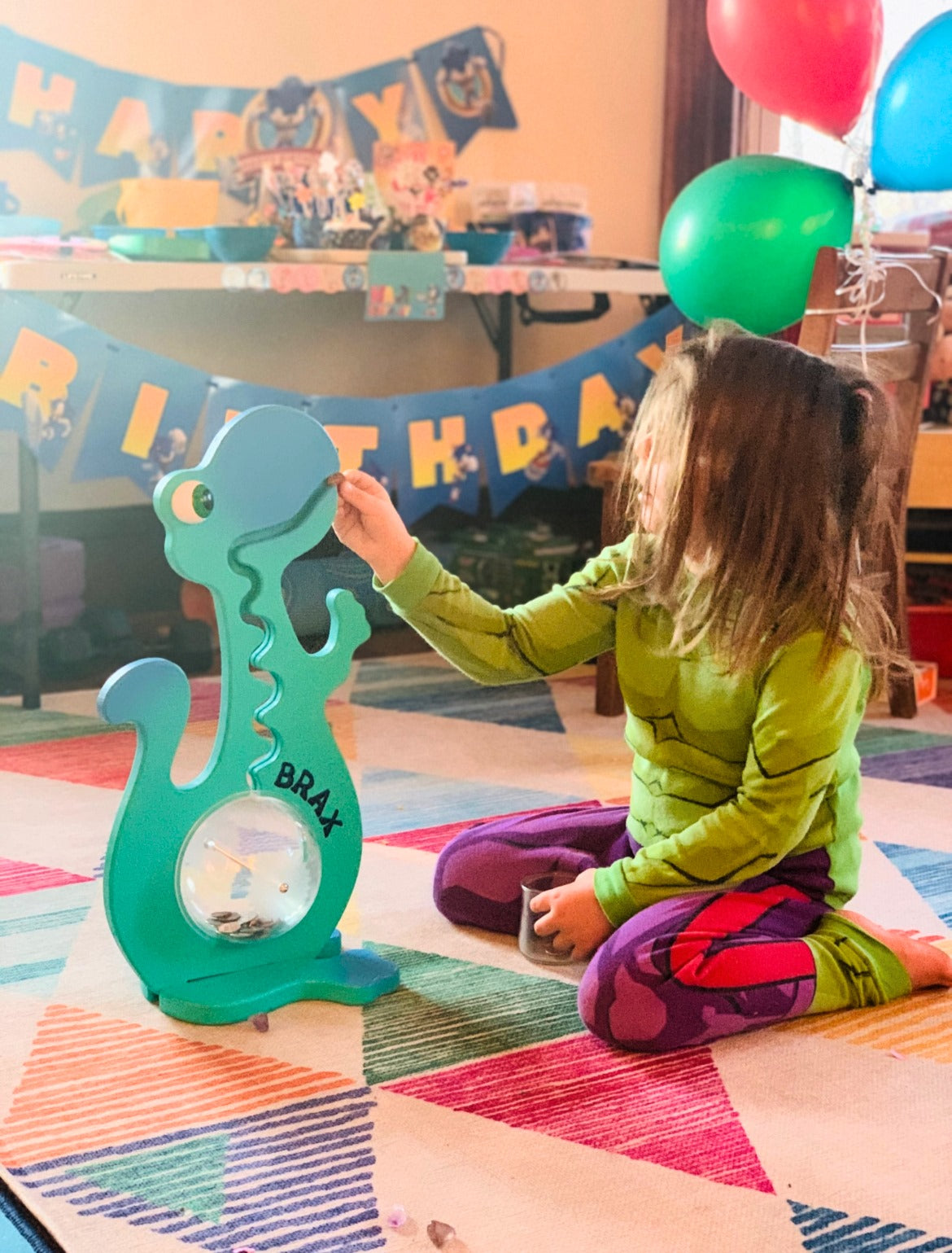 Child beside an aqua dinosaur bank showing personalized lettering at a birthday celebration