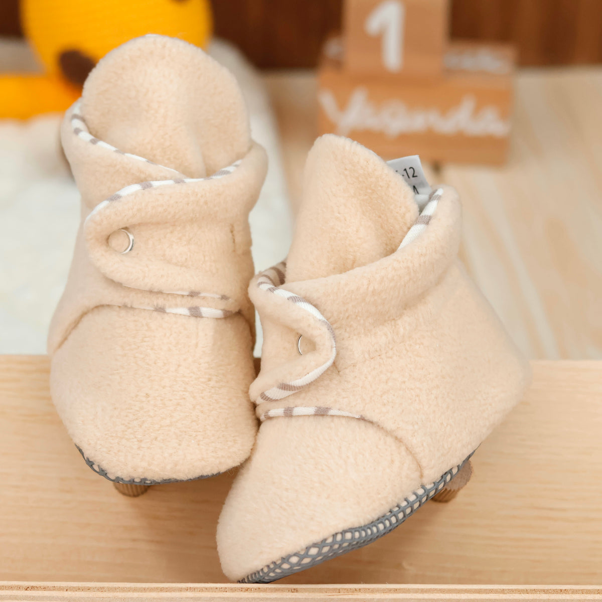 Front view of a pair of cream fleece baby booties with pale striped lining and snap straps.