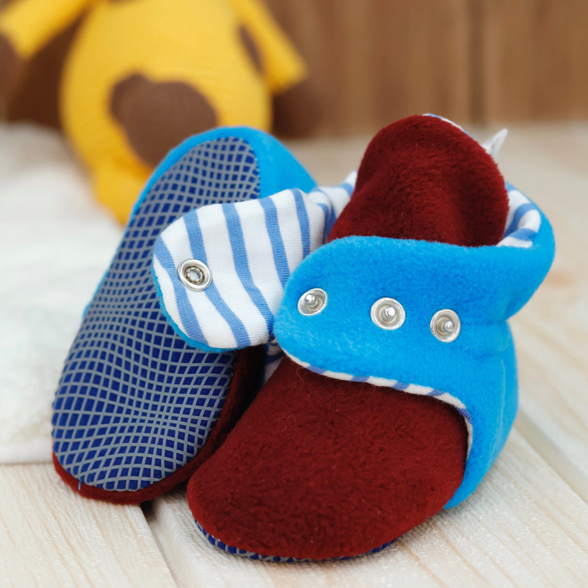 Close-up of blue-and-burgundy fleece baby booties with striped lining and snap straps, showing one sole and an open snap strap.