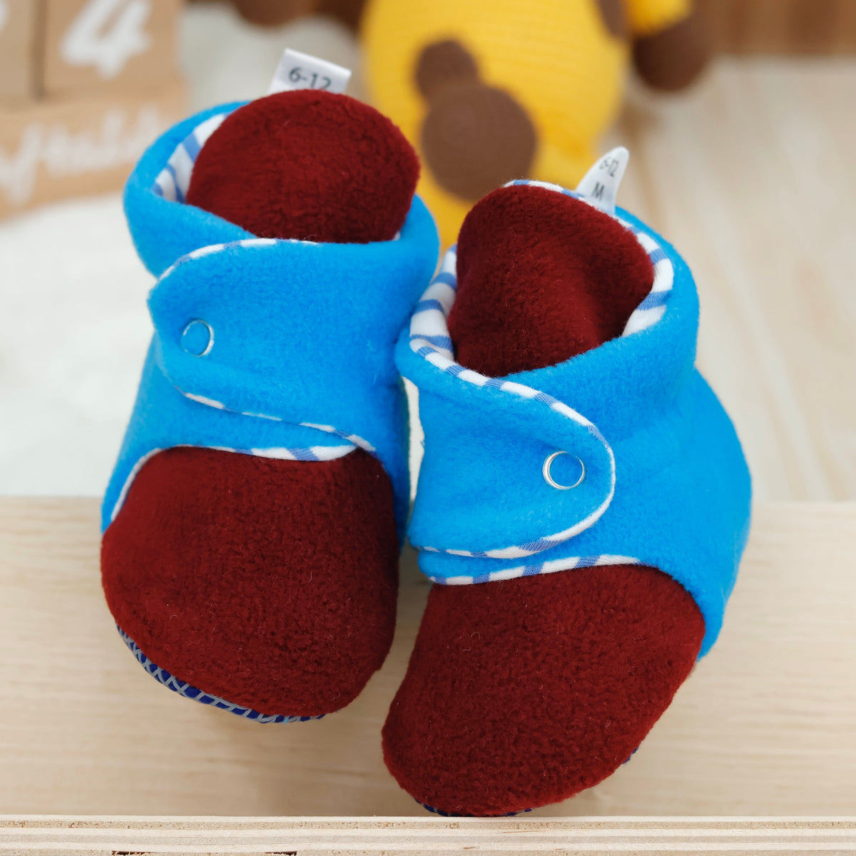 Front view of a pair of blue-and-burgundy fleece baby booties with striped lining and snap straps.