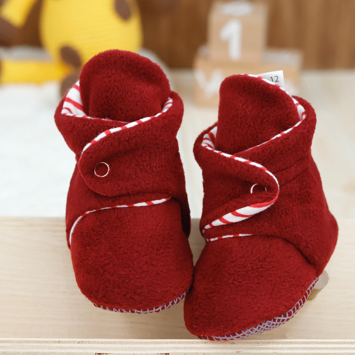 Front view of a pair of burgundy fleece baby booties with striped lining and snap straps.