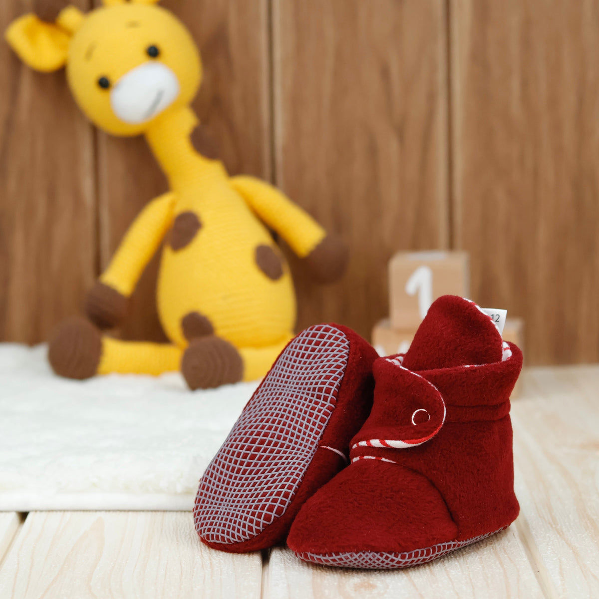 Burgundy fleece baby booties with striped lining and snap straps arranged on a surface with one sole facing the camera.