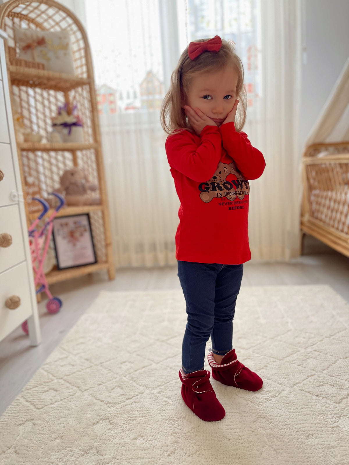 Child standing indoors wearing a red sweater and burgundy booties.