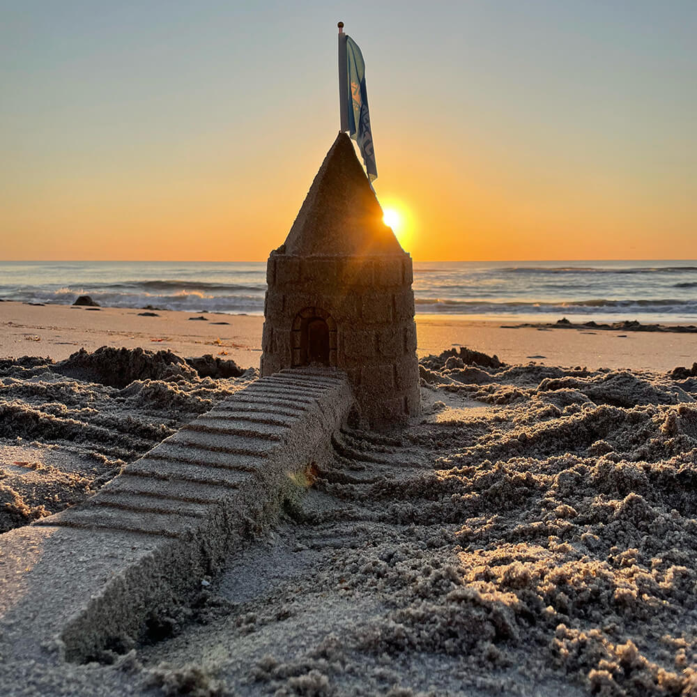Sandcastle tower with a pointed roof, flag and long staircase at sunset.