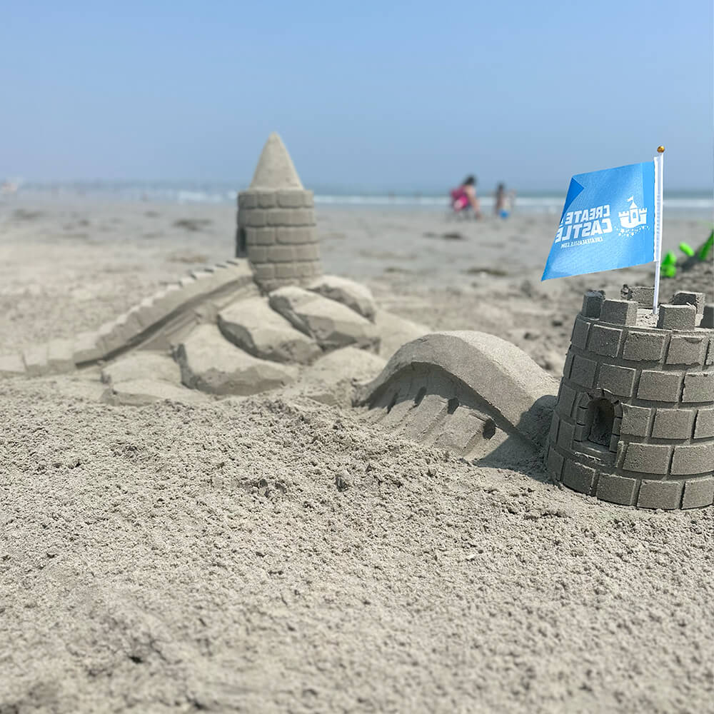 Beach sandcastle scene with a pointed tower, sculpted bridge and foreground turret carrying a blue Create A Castle flag.