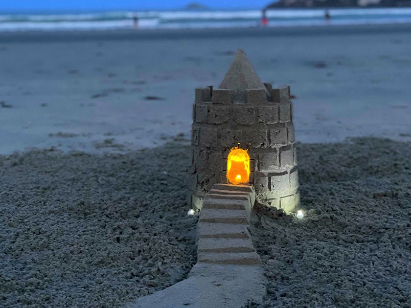 Sandcastle tower and staircase at dusk, lit with an orange glow in the doorway and small lights at its base.