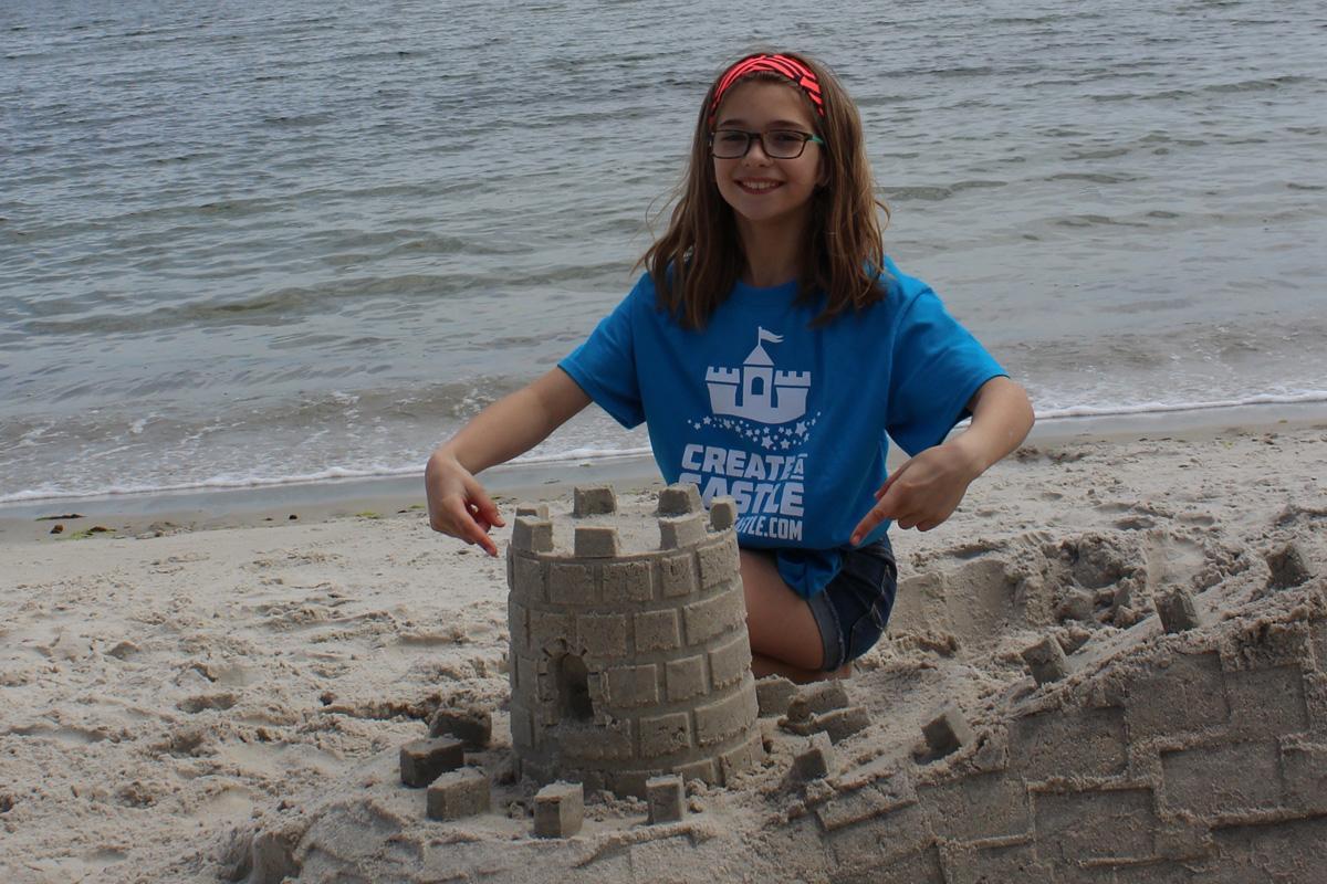 Person in a blue Create A Castle shirt kneeling behind a crenellated sandcastle tower at the shoreline.